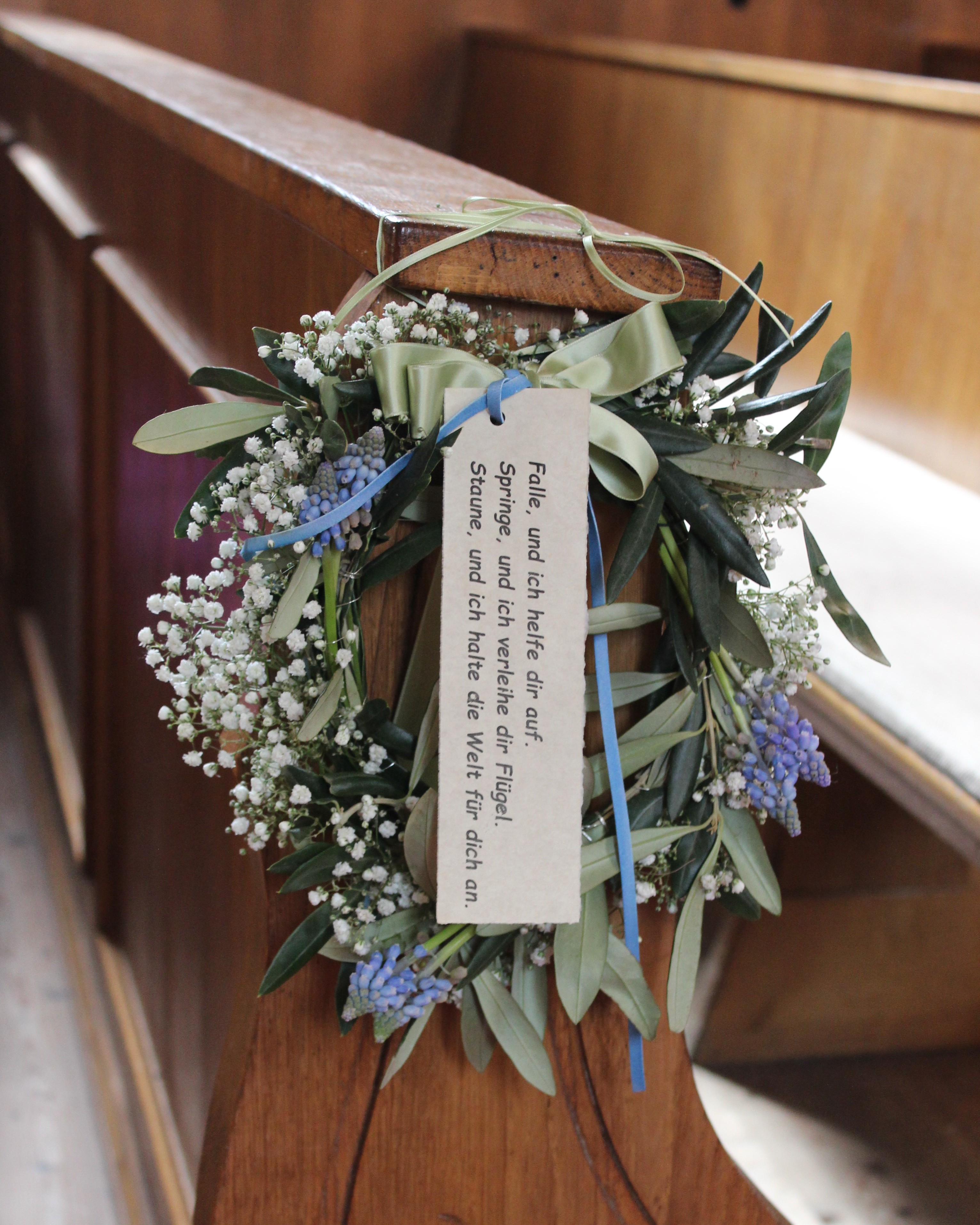 Ein dezenter Kranz aus länglichen Blättern, blauen Blumen, keinen und weißen Blüten und einer großen hellgrünen Schleife