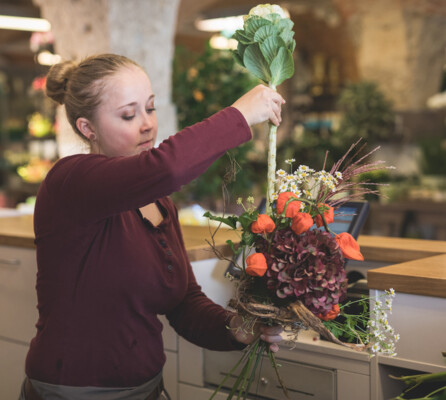 Eine Mitarbeiterin steckt eine langstielige weiße Blume in einen liebevoll zusammengestellten Blumenstrauß aus Blumen in Rot- und Orangetönen